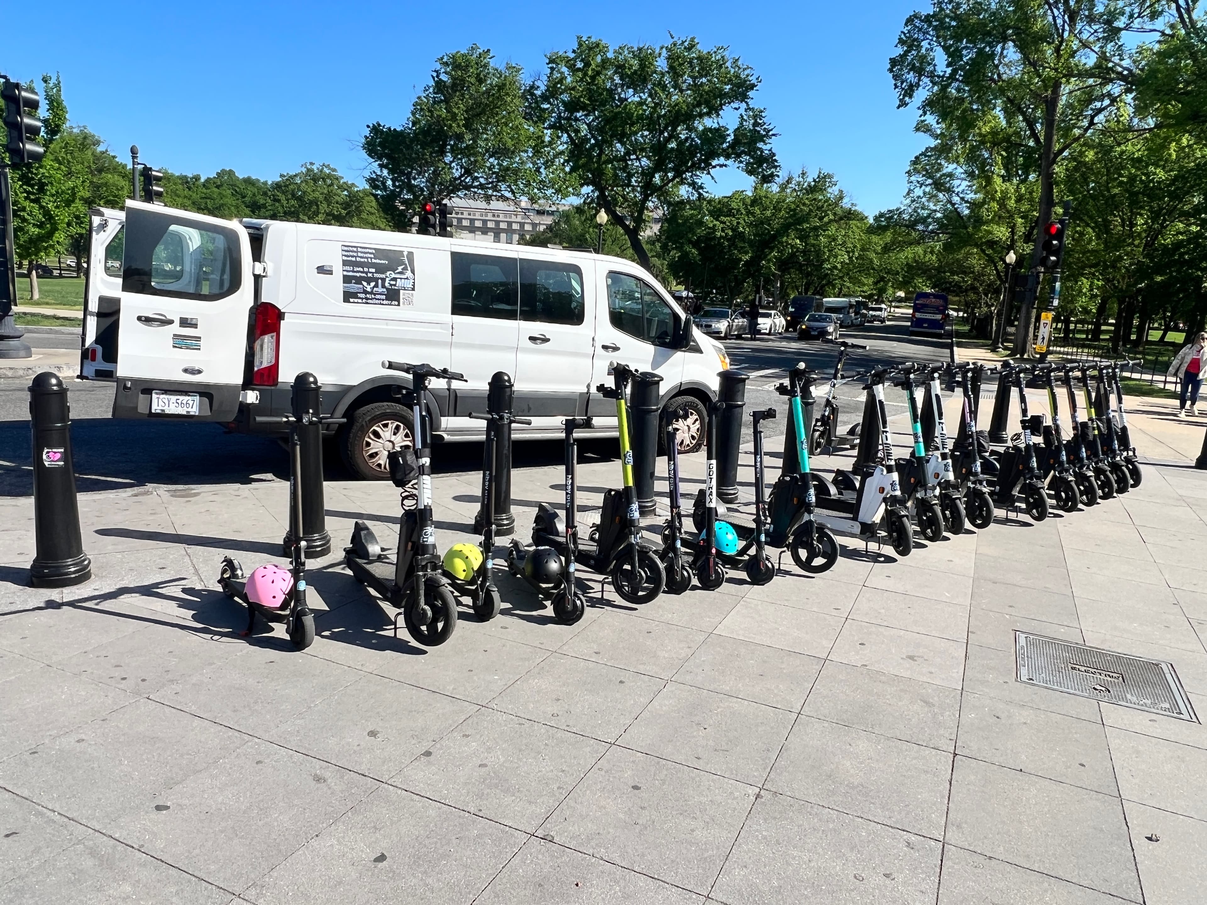 Row of electric scooters with colorful helmets parked beside a white van on a sidewalk.